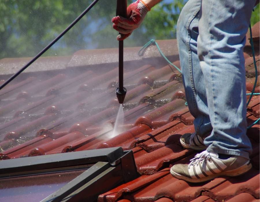 Professional roof cleaner pressure washing a red-tiled residential roof to remove dirt, moss, and algae buildup for improved home maintenance and curb appeal.