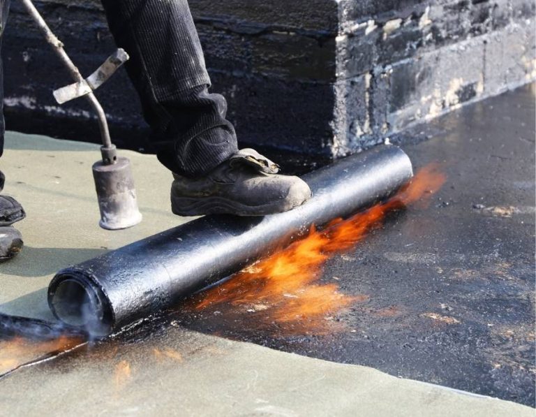 Worker applying bitumen waterproofing membrane with torch-on method for flat roof sealing.